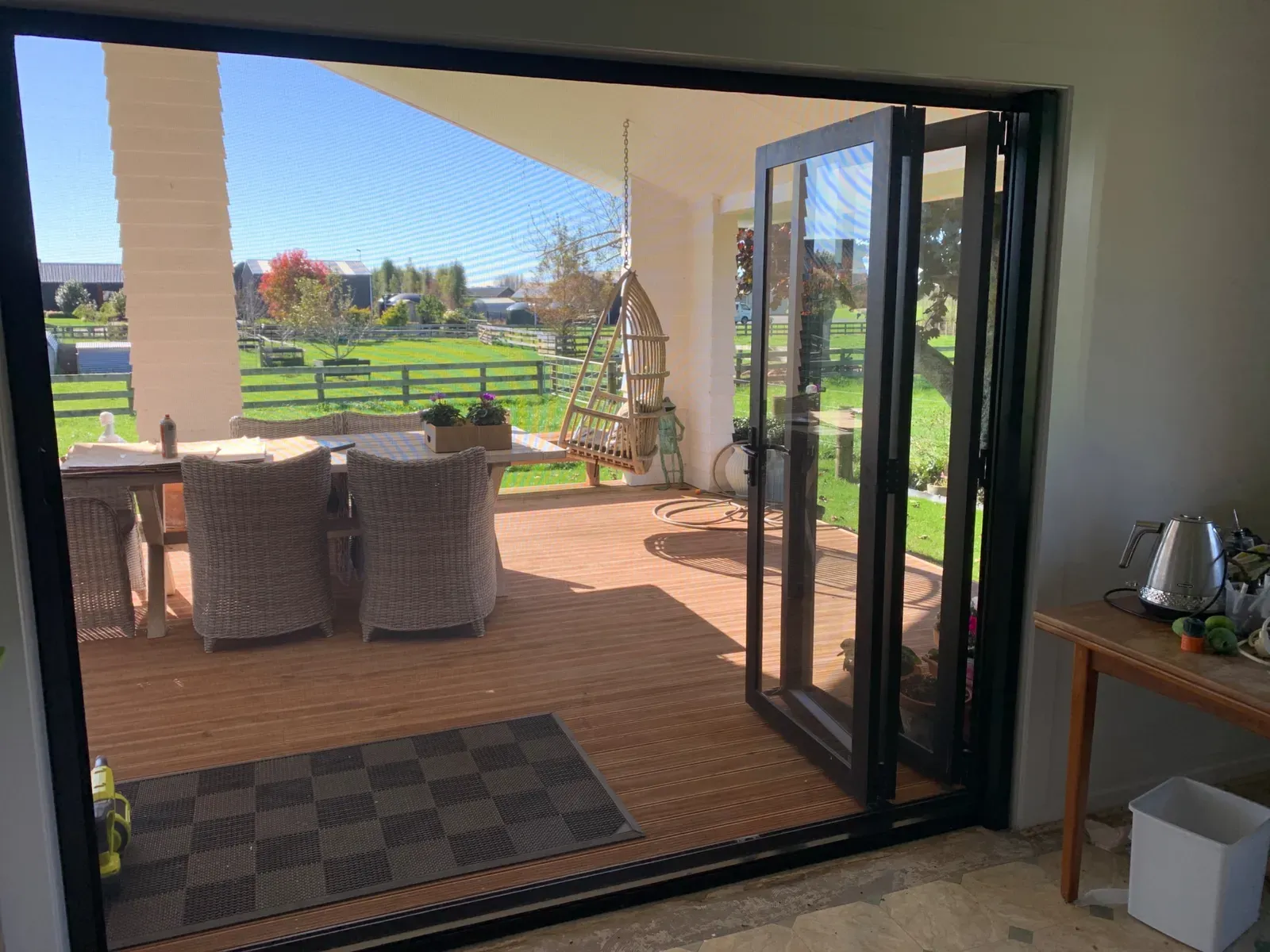 A black retractable Wall Screen drawn across a folded-back bi-fold opening onto a rural deck at Te Kowhai