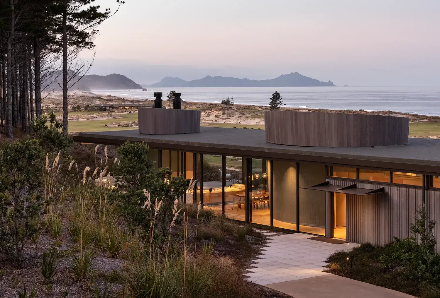 A cedar-clad New Zealand coastal home at dusk overlooking golf links and the sea