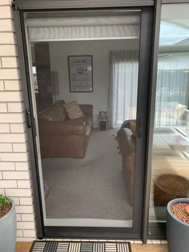 Retractable screen door drawn across a ranch slider at a brick home, leather sofas visible through the mesh