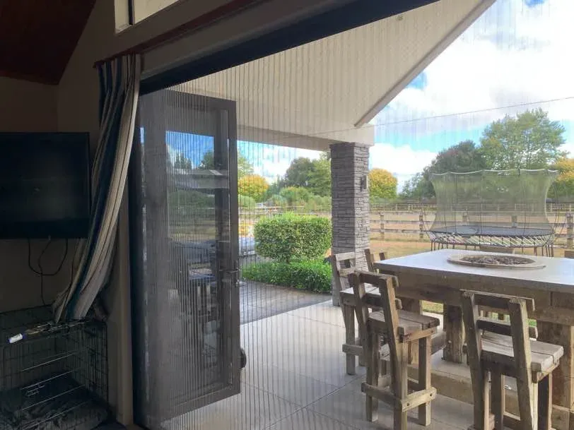 Wide Brio pleated screen across a rural indoor-outdoor opening, an outdoor dining table and paddocks beyond the mesh