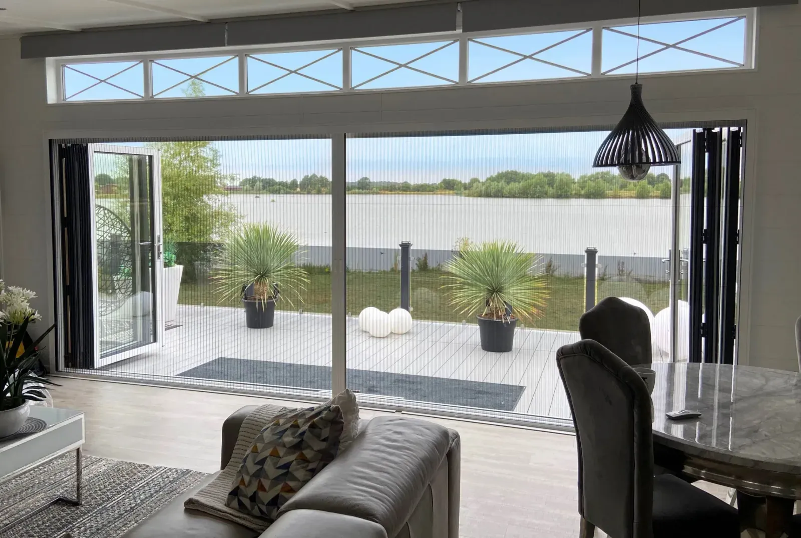 Pleated concertina fly screen drawn across a wide bi-fold opening, doors folded back onto a lakeside deck
