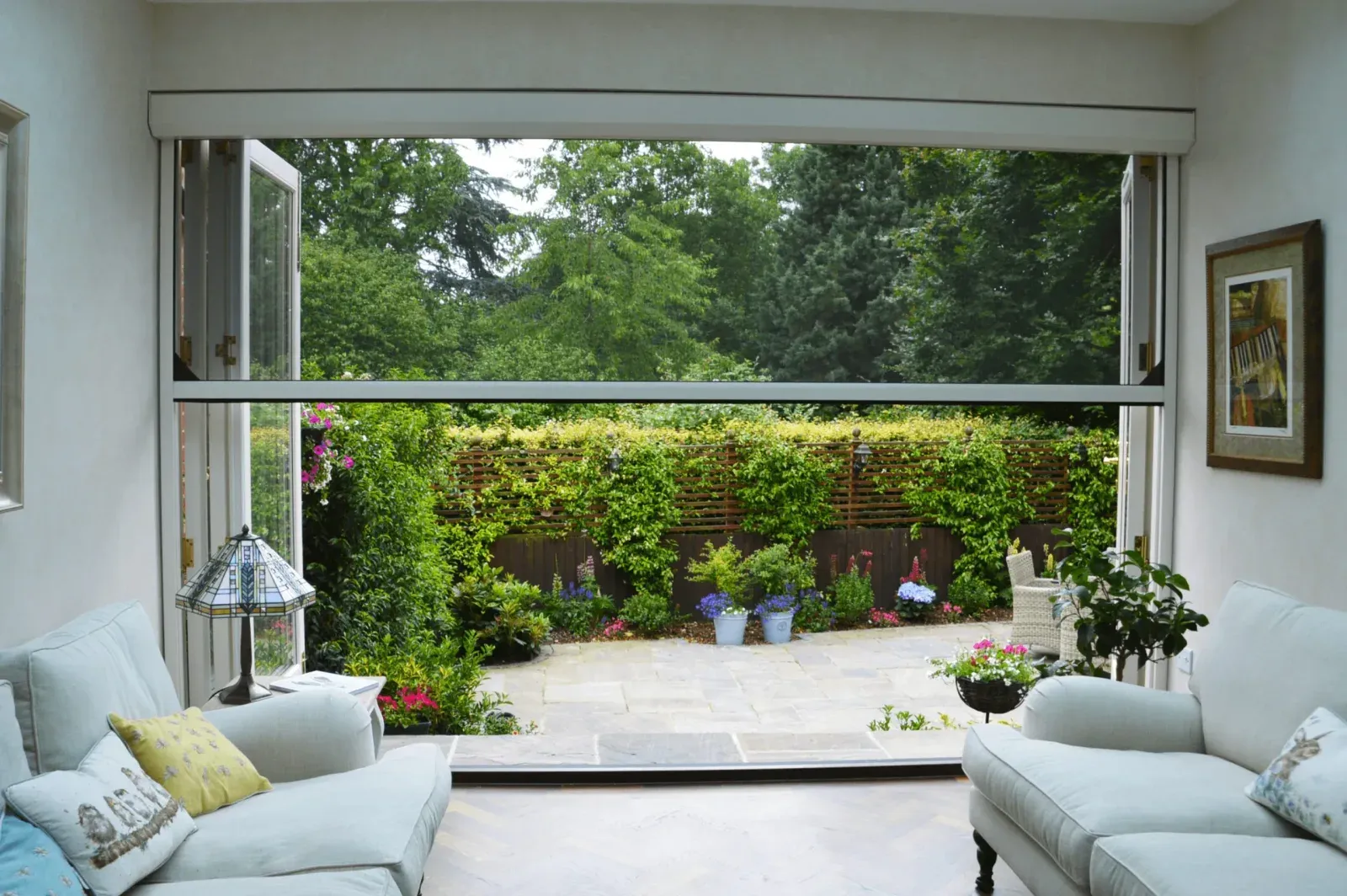 Clear-view insect-mesh outdoor screen retracted above a wide open patio doorway, seen from the lounge inside looking to a garden