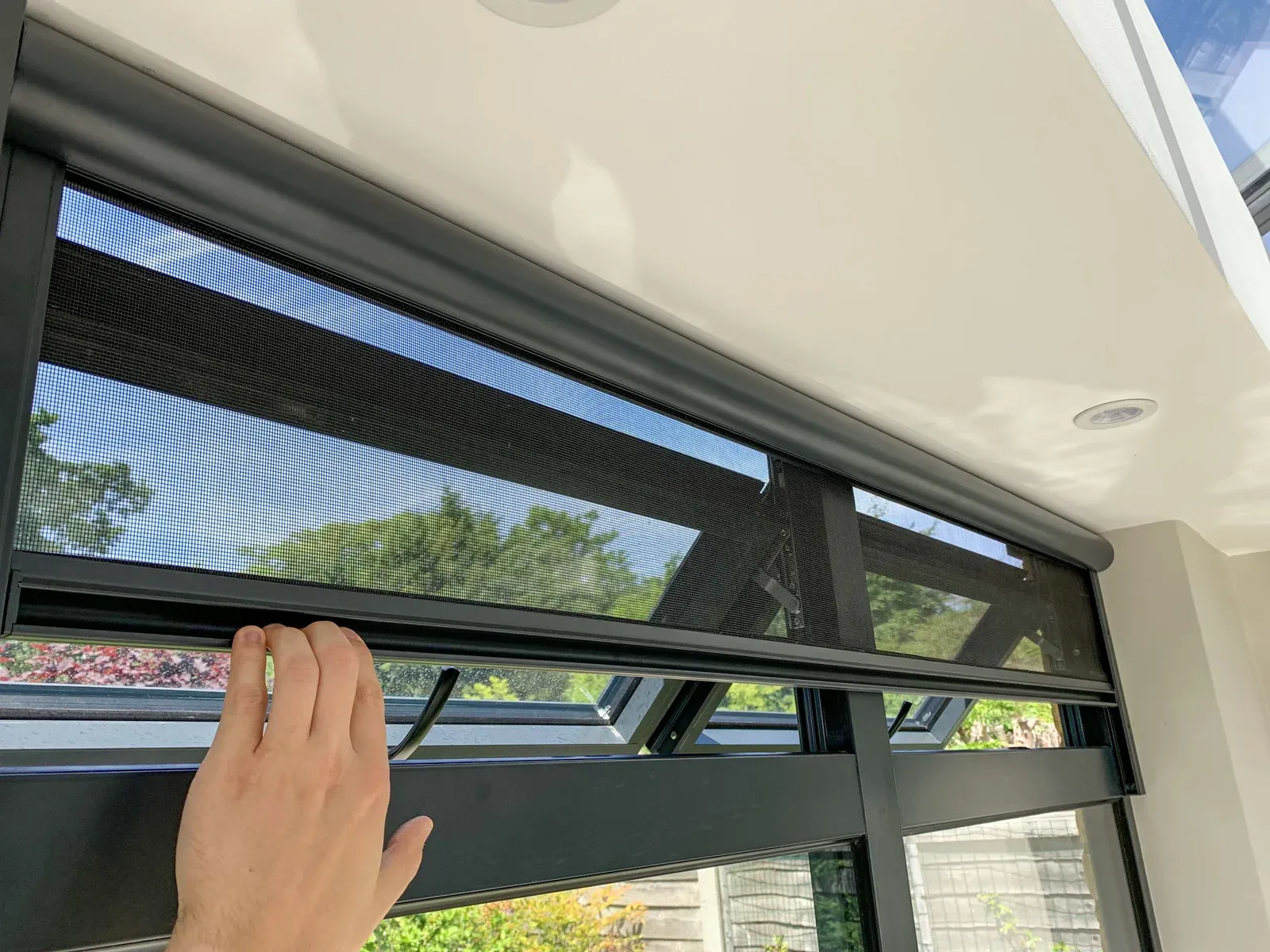 Black insect mesh Serene retractable screen over a black-framed double awning window, hand on the bottom bar