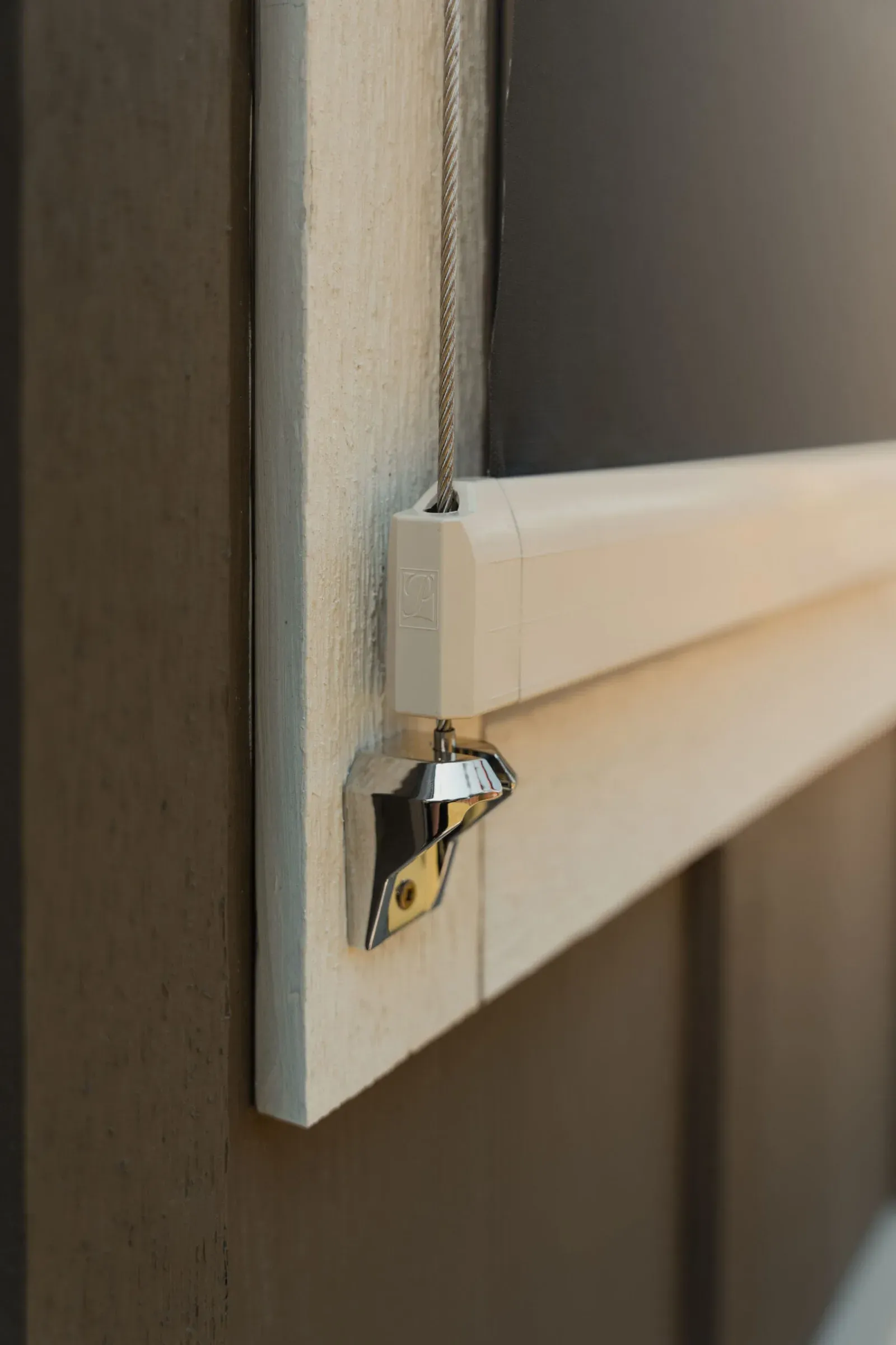 Close-up of an outdoor screen's branded aluminium bottom-bar end cap, stainless side cable and a polished chrome tie-down cleat on a timber jamb