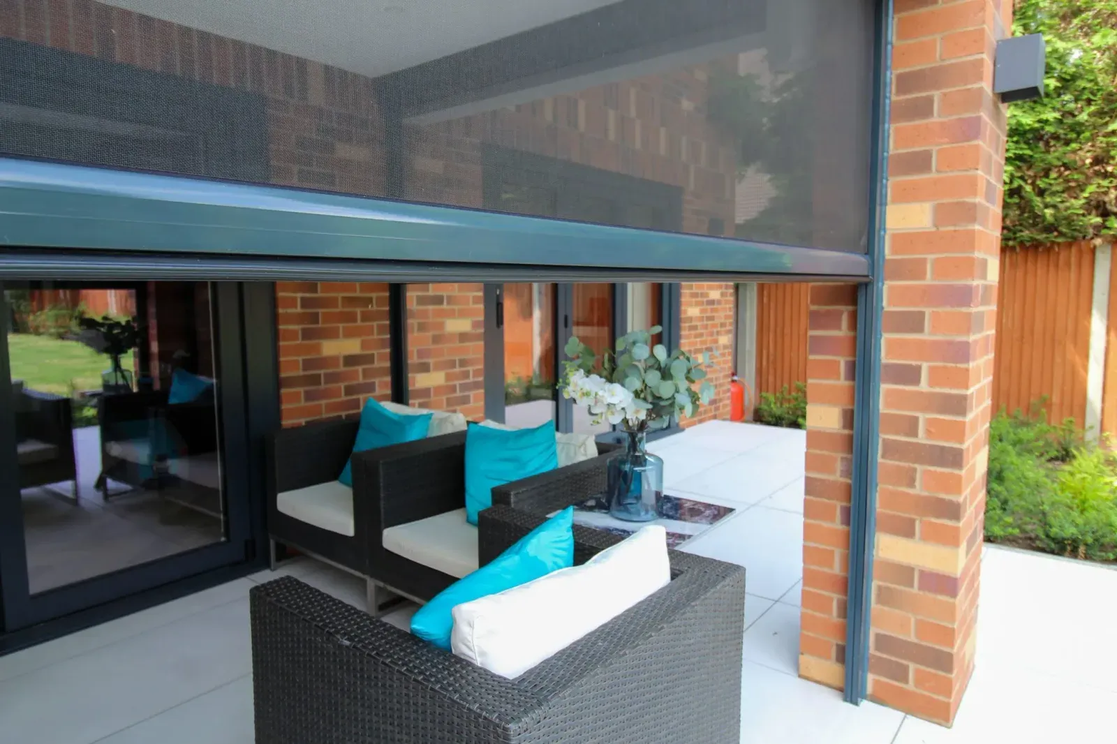 Black solar-mesh outdoor blind dropped from the soffit over a covered brick patio with lounge seating