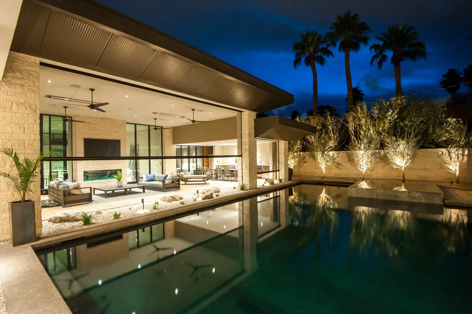 Motorised outdoor screens retracted across the large openings of a covered poolside outdoor room lit at dusk