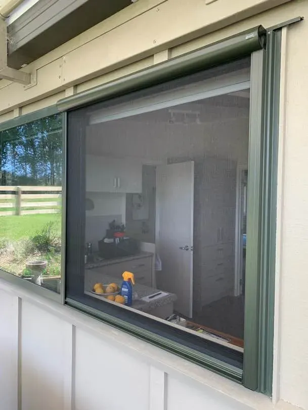 Green-framed retractable roller screen fitted over a large window, viewed from outside against a rural paddock