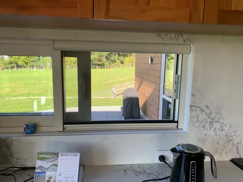 Retractable window screen cassette mounted above a kitchen slider window, overlooking a lawn and patio