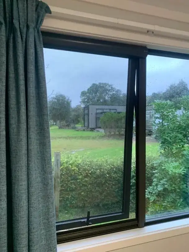 A retractable insect screen drawn across an open awning window at Opoutere, looking over a grassy section to a small cabin