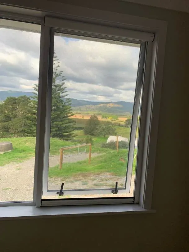 Retractable insect screen over an awning window, looking out to a farm gate, paddocks and distant ranges