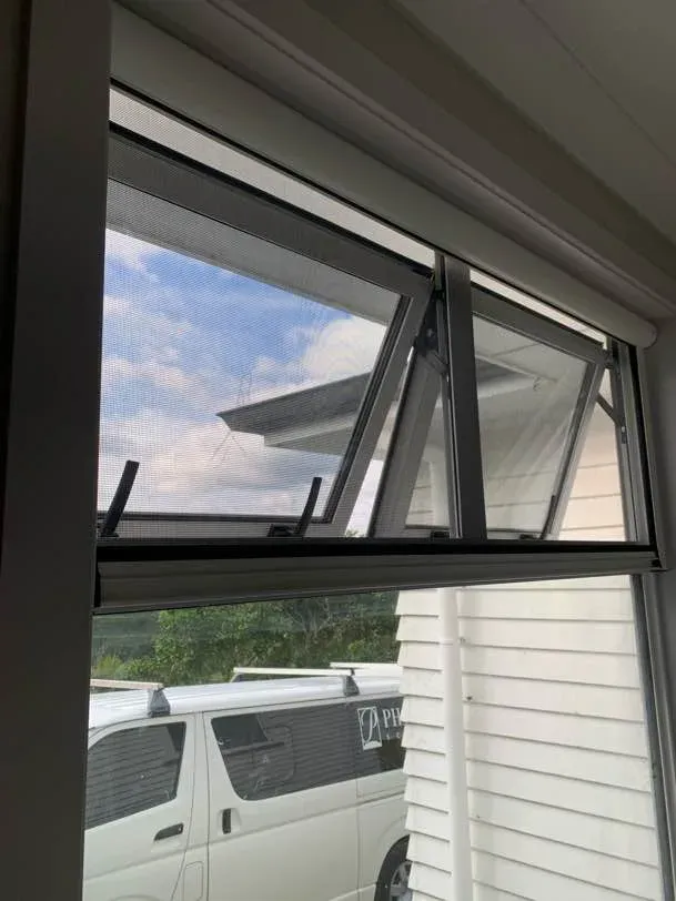 Black-framed retractable insect screen across an awning window opened outward to a bright sky