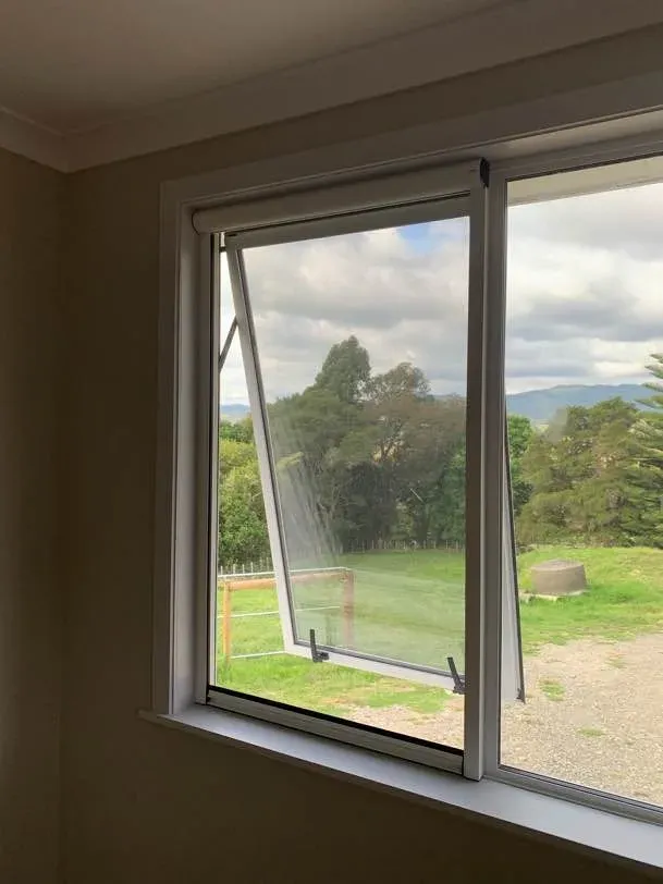 Corner window with an awning sash open behind a retractable insect screen, overlooking a green paddock