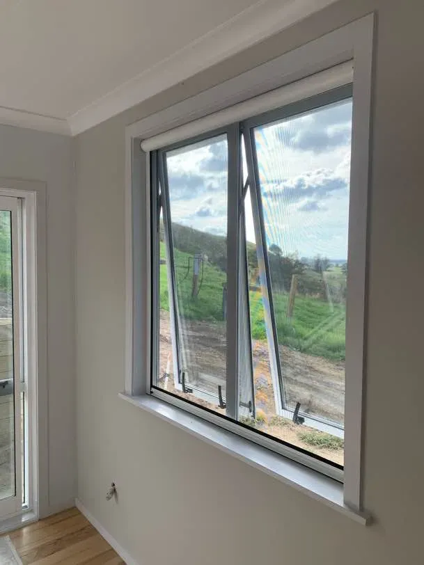 Two awning windows tilted open behind fitted retractable insect screens, looking out over rolling farmland