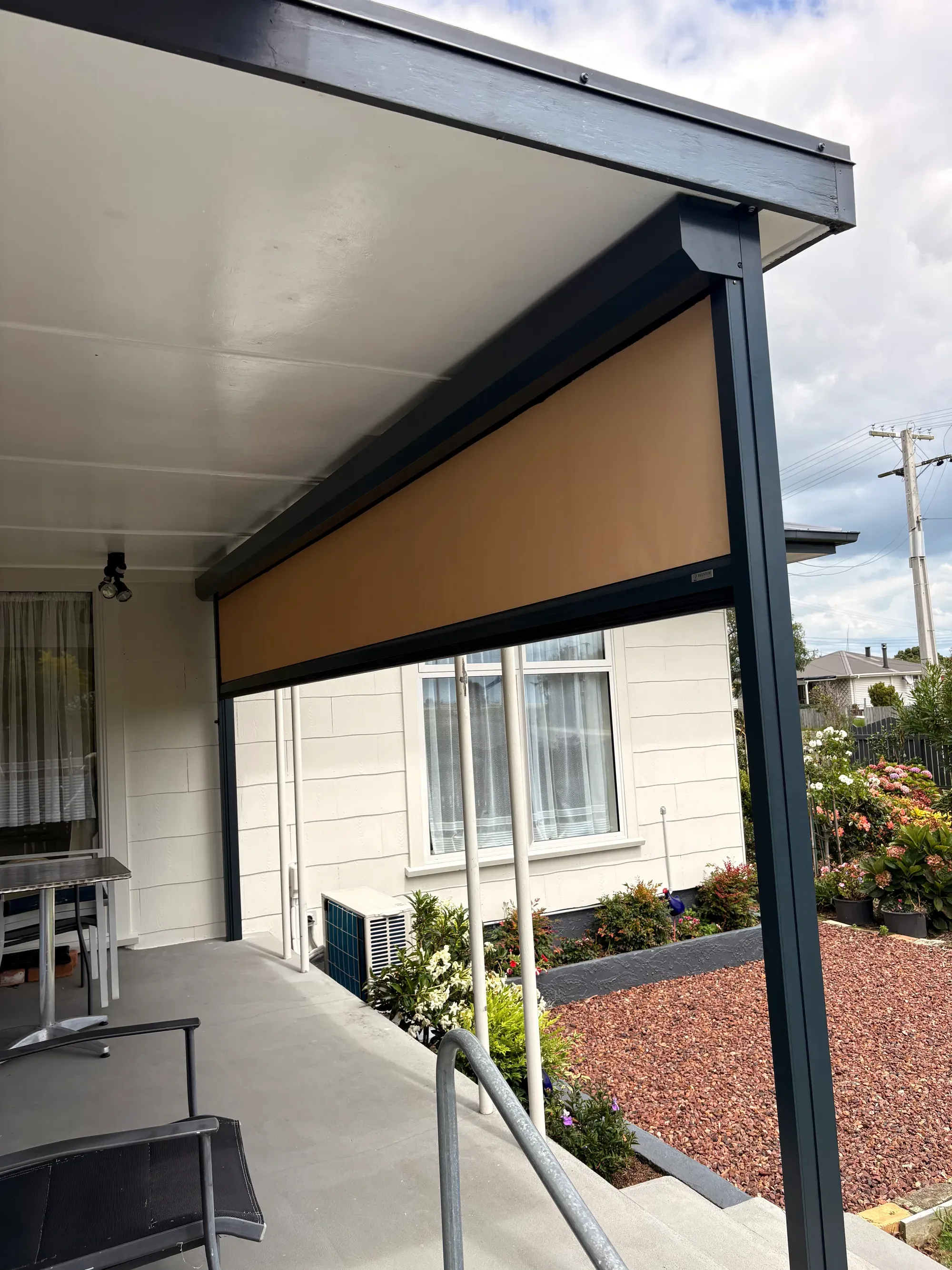 A tan outdoor blind part-lowered under a dark-framed patio roof over a residential deck, house and garden beyond