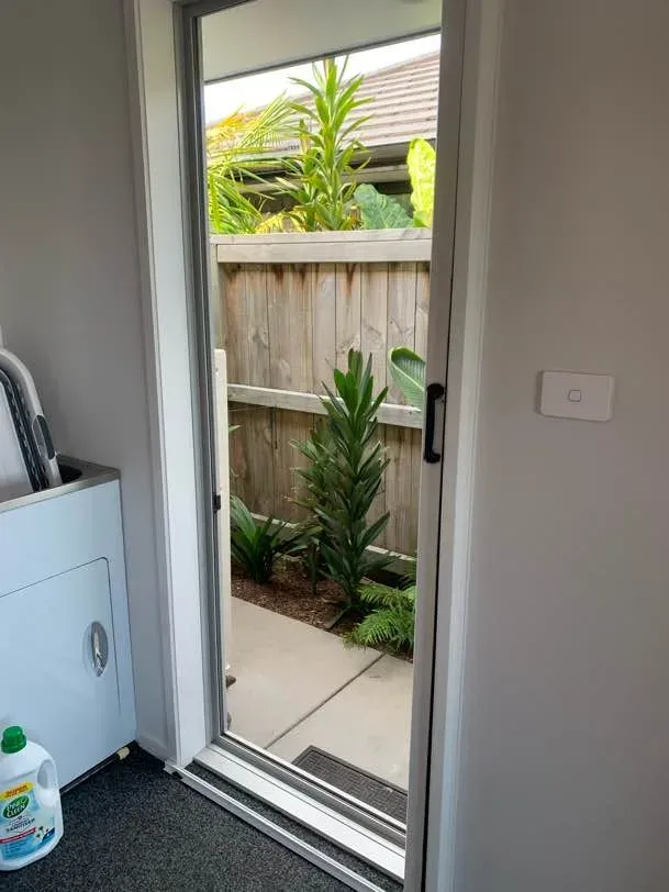 White-framed retractable insect screen door across an open doorway leading out to a fenced garden with subtropical planting