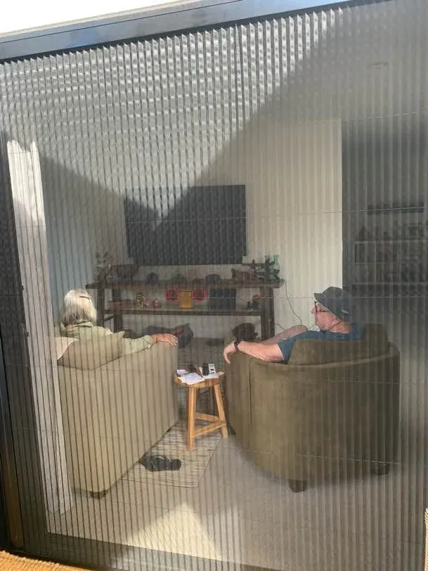 Two people relaxing in armchairs, seen from a sunny deck through a retractable pleated screen drawn across a wide living-room opening