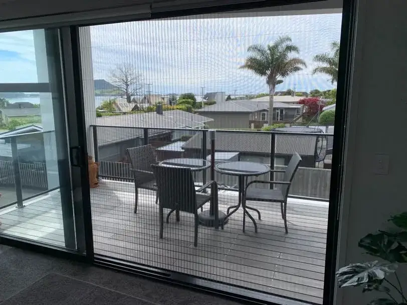Retractable pleated insect screen across a sliding door, viewed from inside toward a deck with Tauranga harbour views