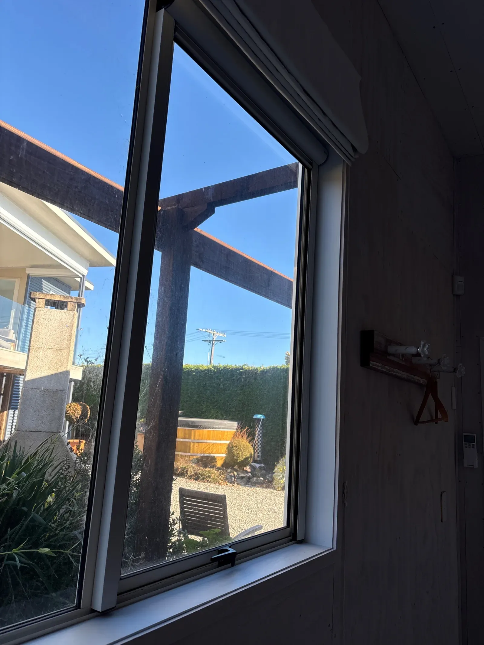 Awning window with a dark blockout roller part raised, looking out to a gravel courtyard with a wooden hot tub