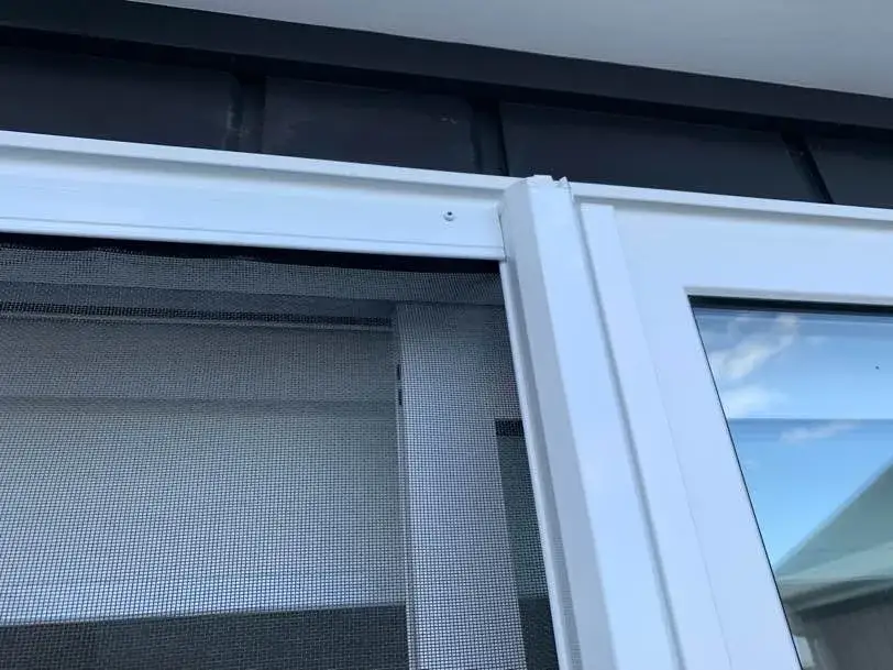 Close-up of the mesh and white top track of a retractable insect screen fitted to the white joinery of a dark-clad Te Puke home, blue sky reflected in the glass beside it