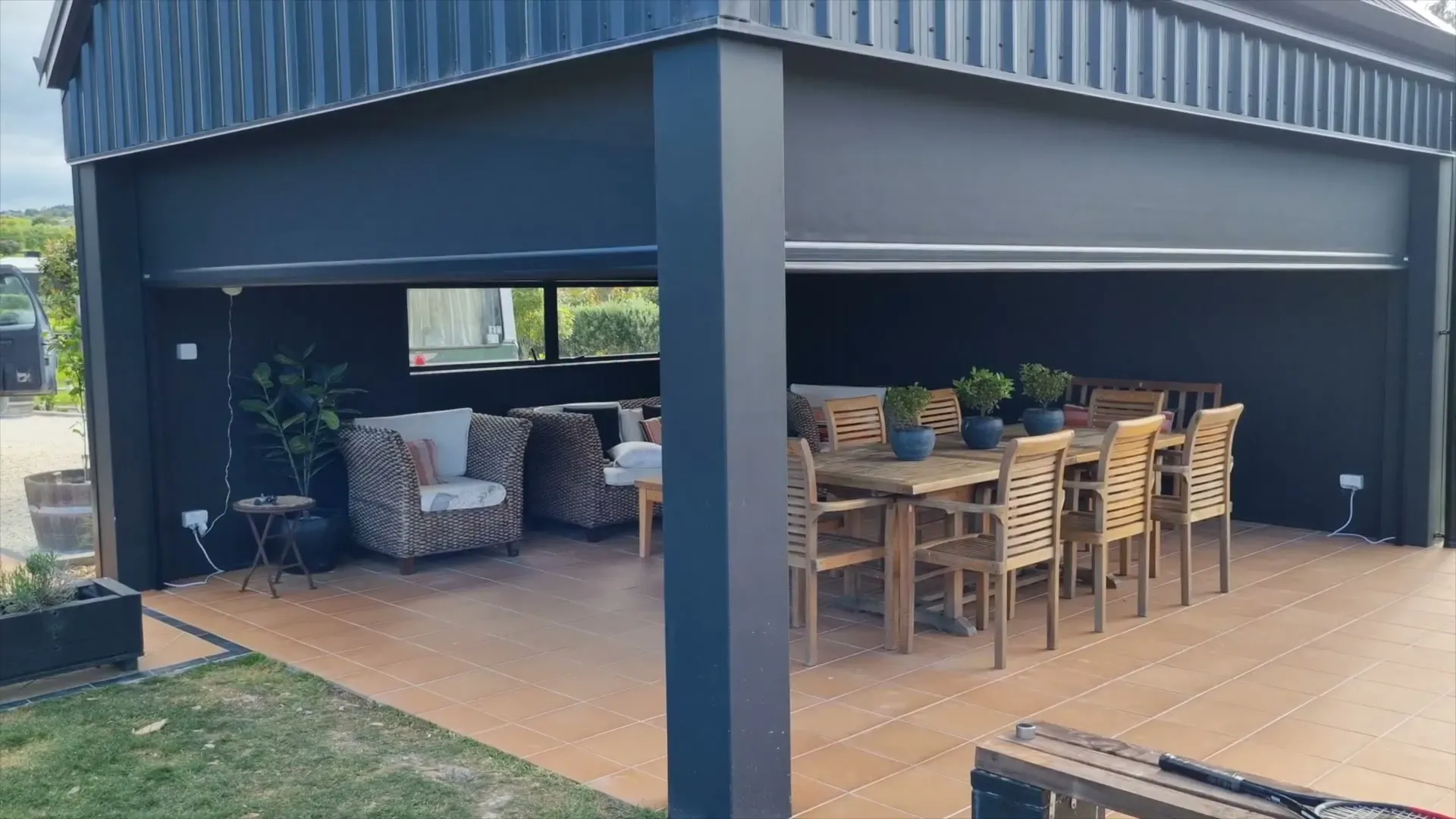 Executive motorised screens part-lowered around a covered outdoor entertaining area with a dining table and lounge
