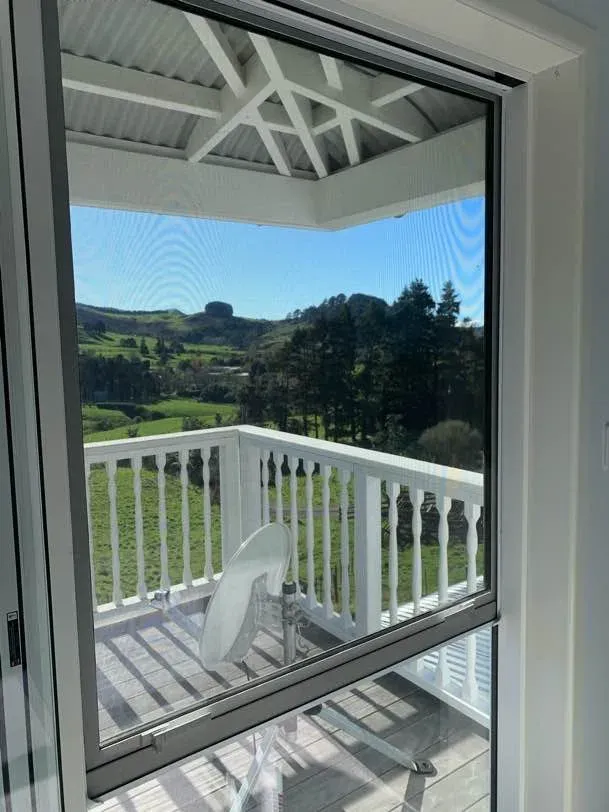 Looking through a mesh-screened window to a white verandah balustrade and green hill country