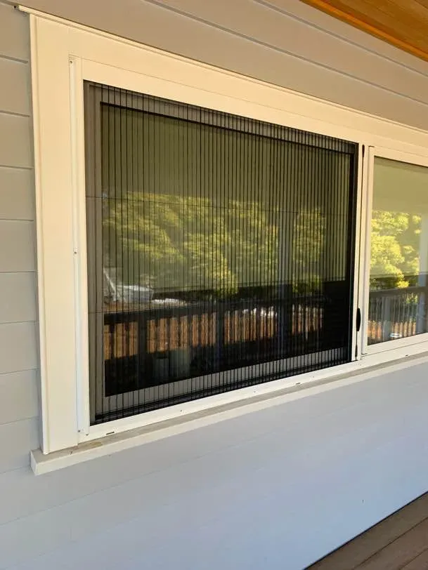 Black Brio pleated screen drawn across a wide sliding window on a grey weatherboard home, deck balustrade beyond