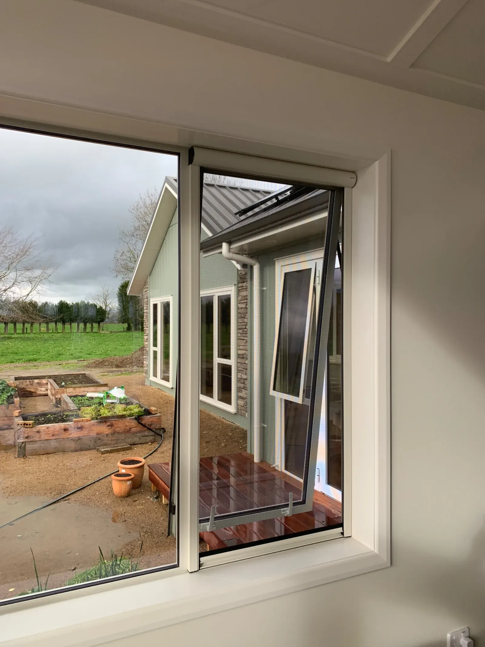 White Serene retractable screen drawn down over an open awning window, looking out to a rural garden and paddock