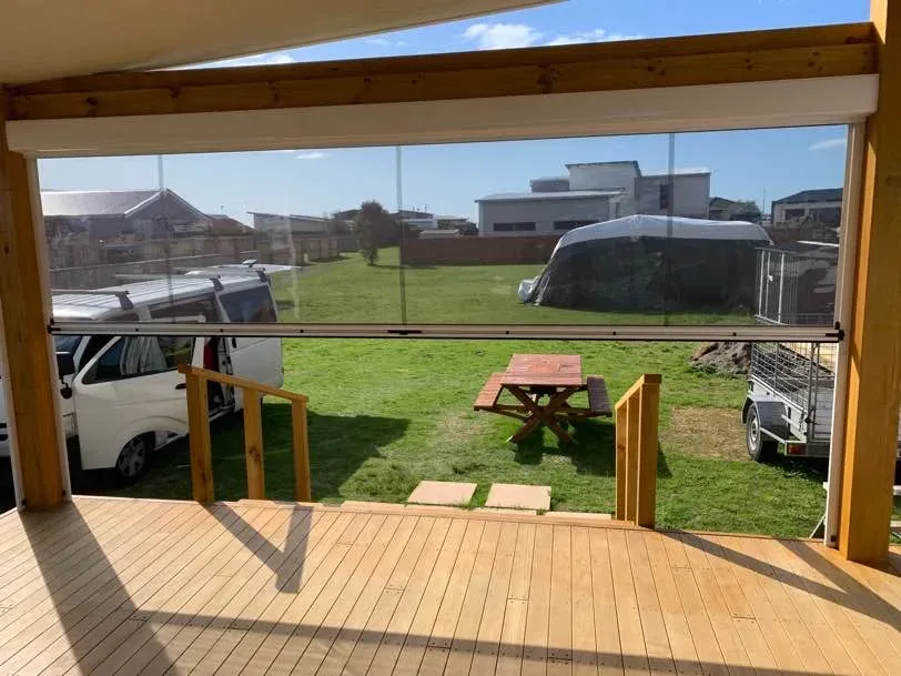 Clear PVC outdoor blind drawn down across a covered timber deck opening, with lawn visible through it